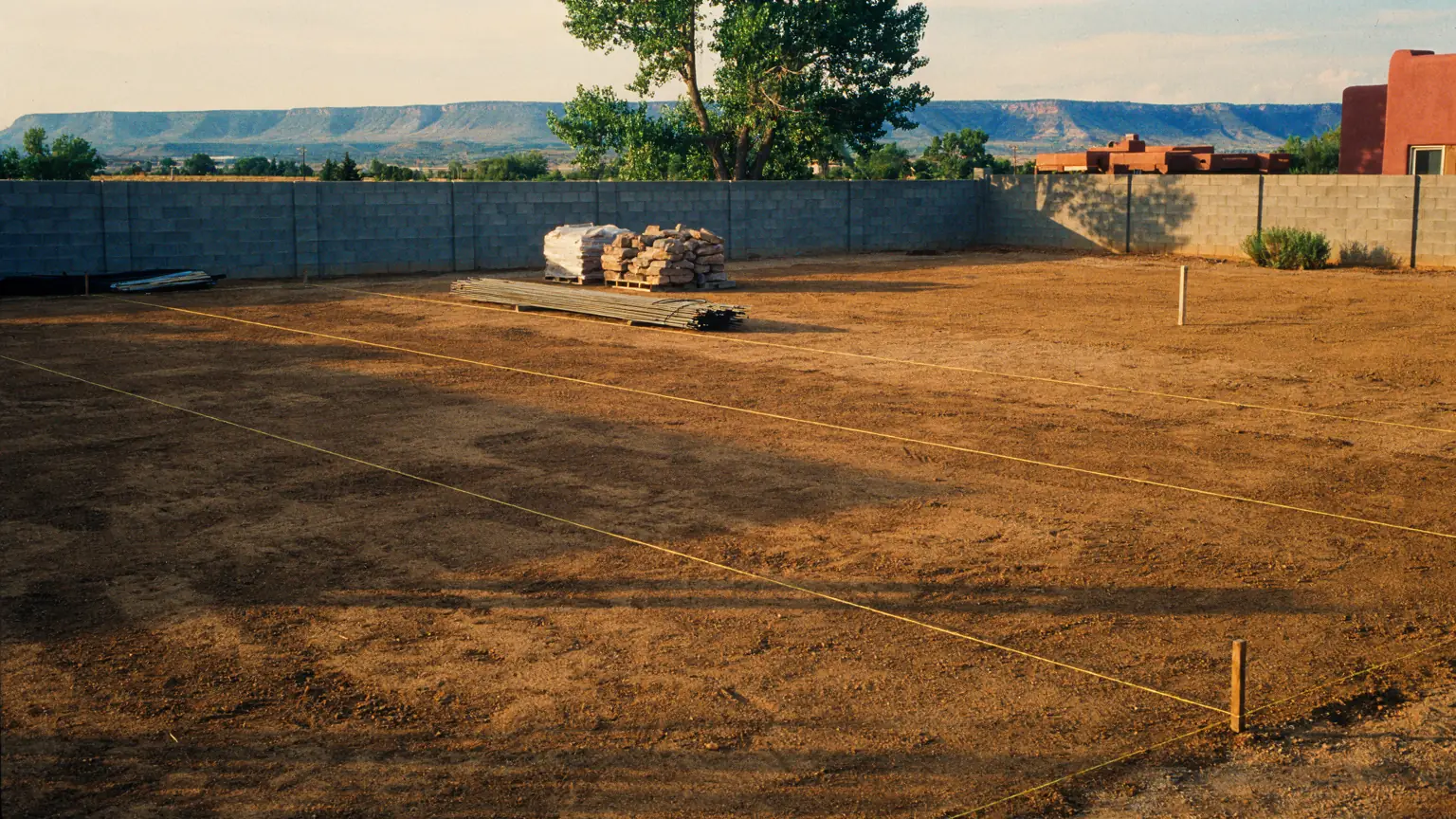 A Rio Grande valley yard mid-conversion: graded ground with taut string lines, stacked steel edging and a pallet of rock, block wall and one cottonwood behind